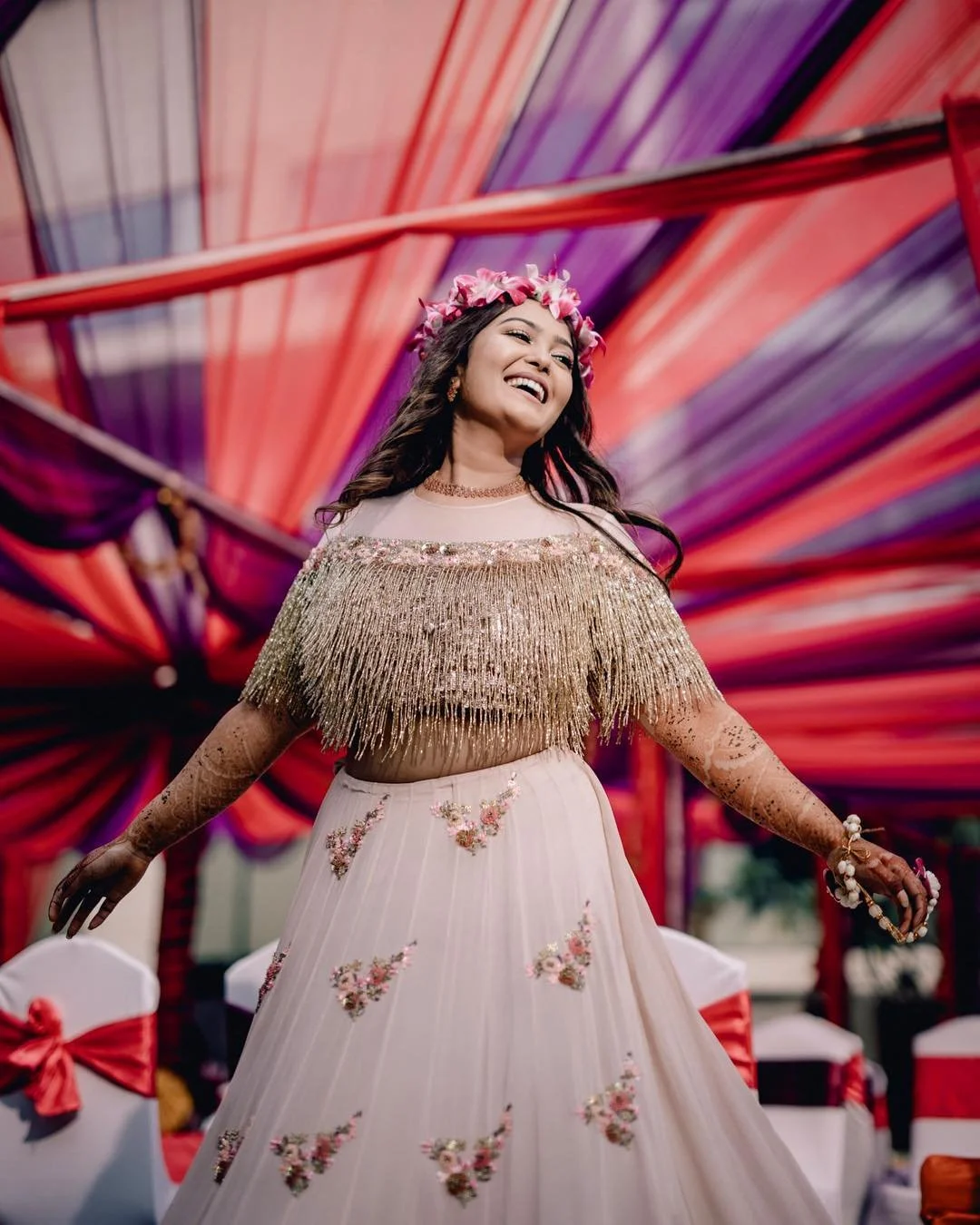 A woman in a traditional attire, wearing a flower crown, smiling at a vibrant outdoor event with a colorful fabric canopy in the background.