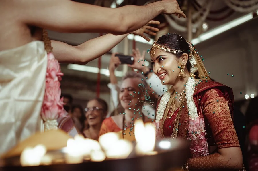 An Indian bride wearing traditional jewelry and a maroon saree, smiling as water is poured over her head during a wedding ceremony, surrounded by family members and friends.