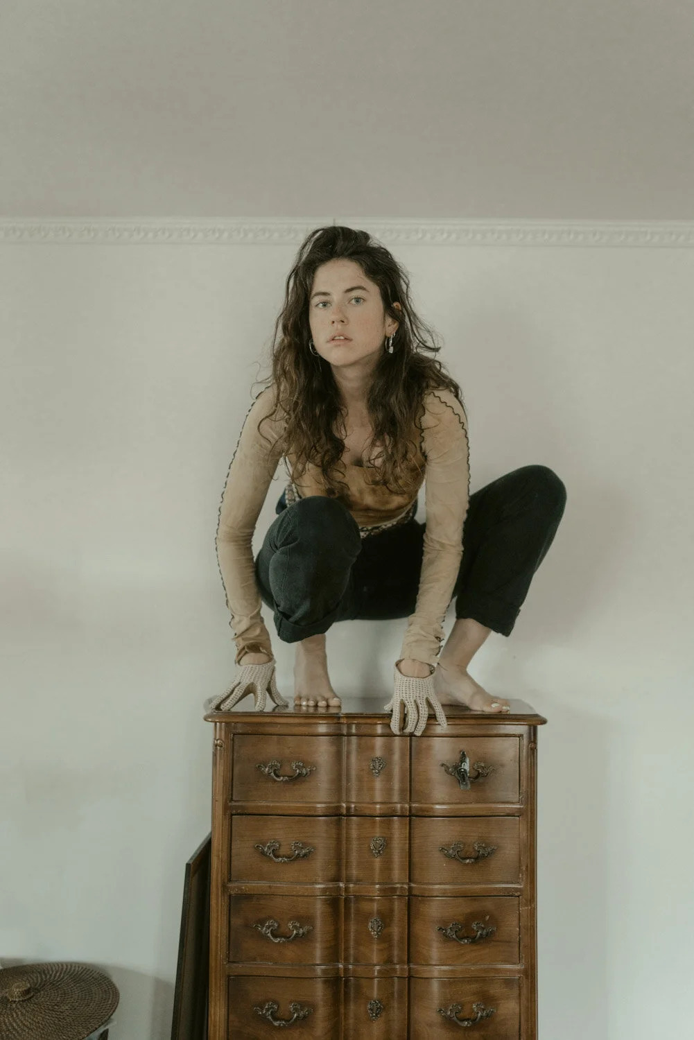 A woman with curly hair crouching on top of a wooden dresser, wearing a beige long-sleeve top, black pants, and gloves, indoors against a plain wall.
