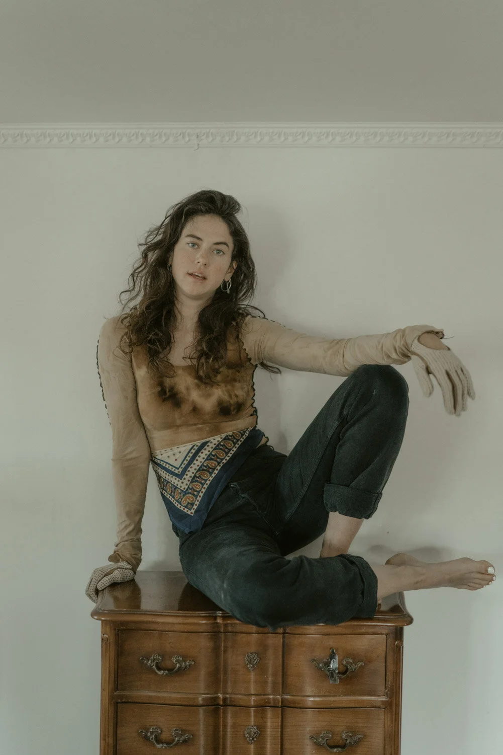 A woman with curly brown hair wearing a tan long-sleeve shirt, black pants, and beige gloves, sitting on top of a wooden dresser against a plain white wall.