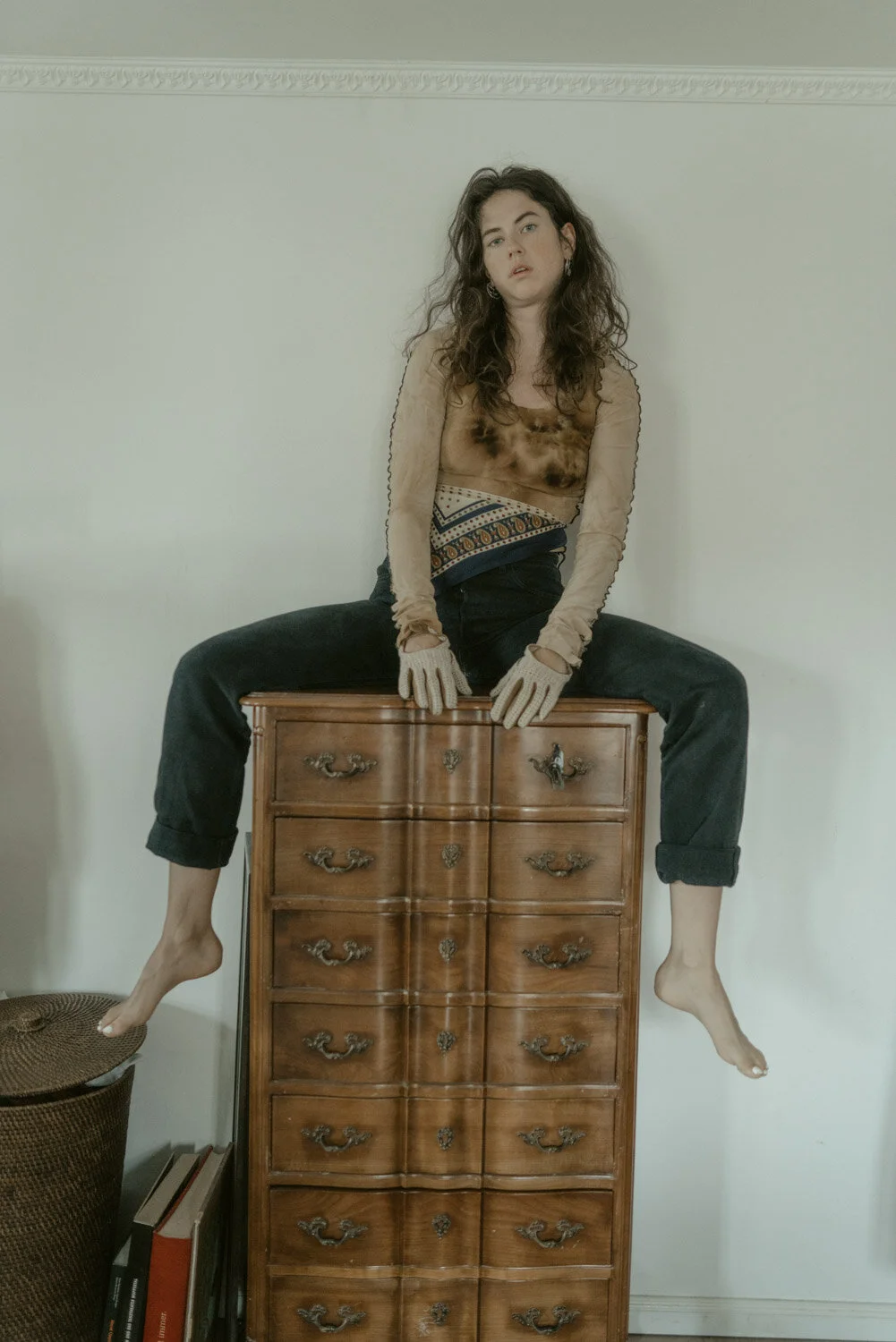 A woman with long curly hair sitting on top of a wooden dresser with her legs spread apart and hanging down, wearing a beige top, black pants, and gloves, with a neutral expression on her face.
