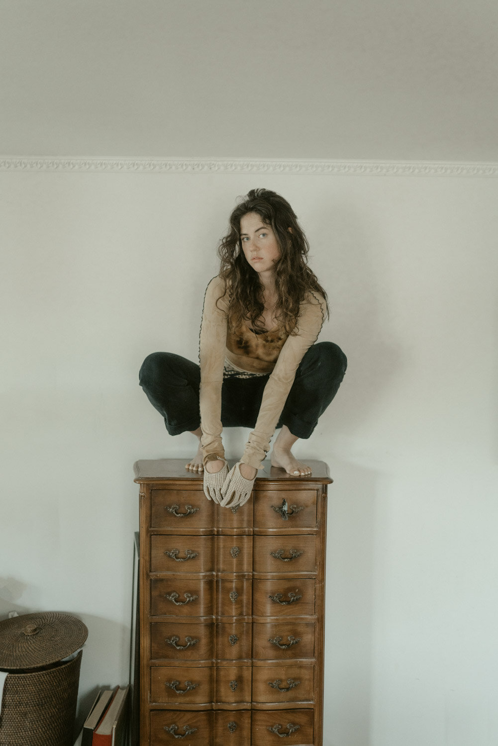 Young woman with curly brown hair crouches on top of a wooden dresser, barefoot, wearing long-sleeve beige shirt, black pants, and gloves, looking at the camera in a room with a plain beige wall.