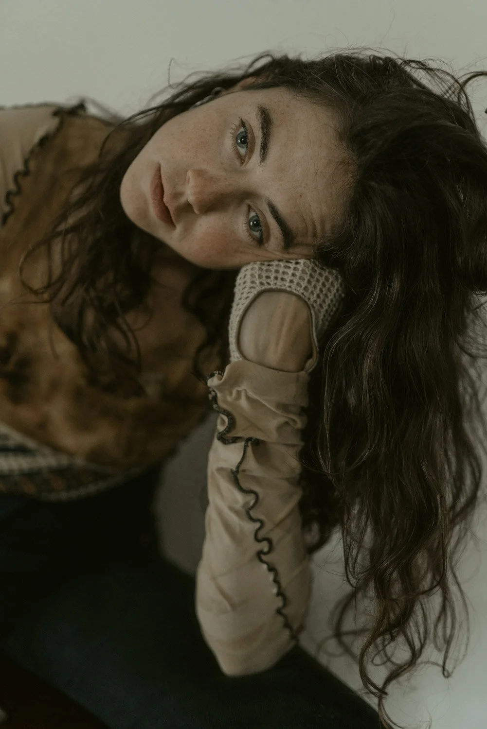 A woman with long, dark, curly hair lying on her side, resting her head on her hand, looking directly at the camera with a contemplative expression.