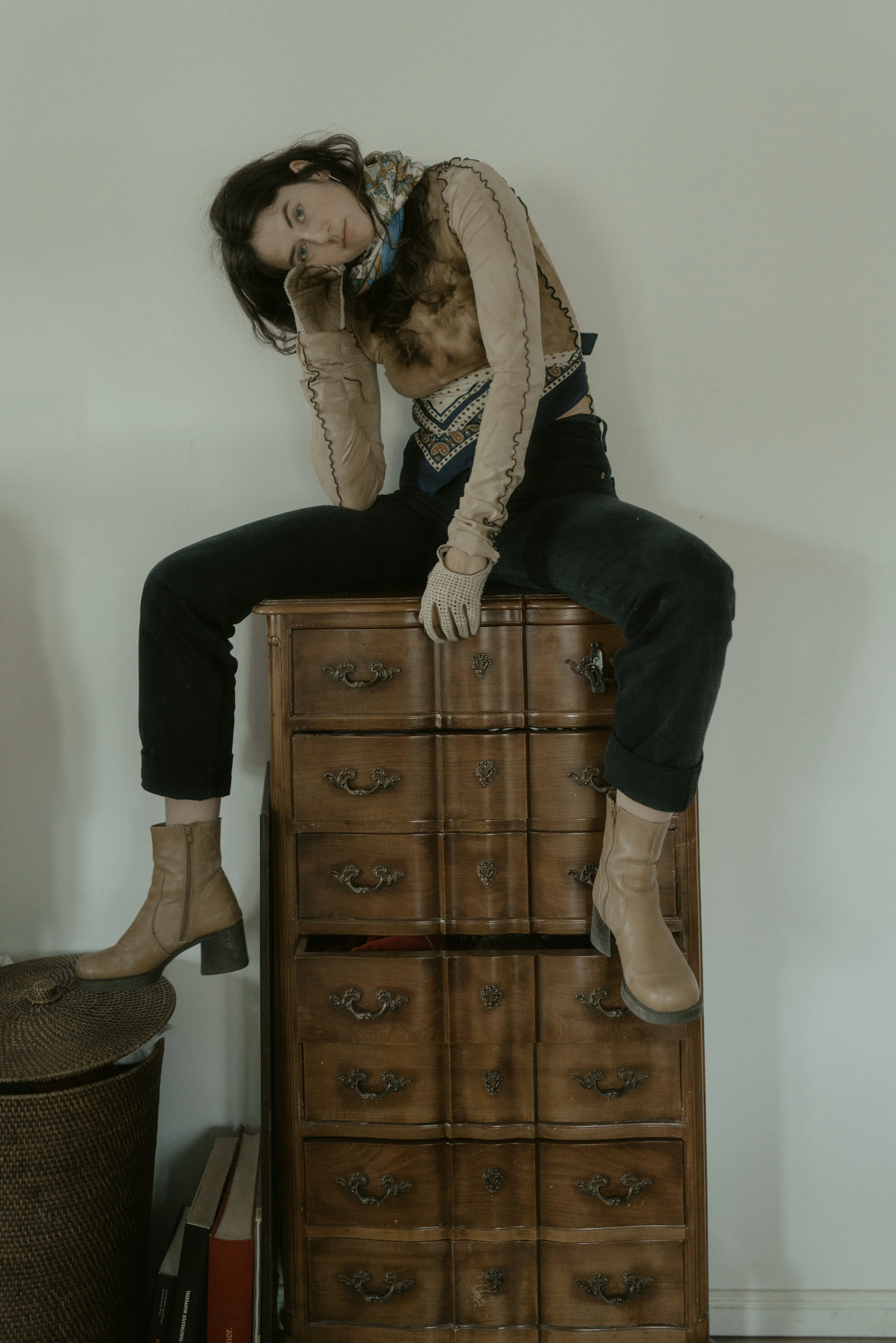 A woman sitting on top of a wooden dresser with one hand on her forehead and the other hand resting on her lap, wearing beige boots, black pants, a beige patterned top, and a multicolored scarf.