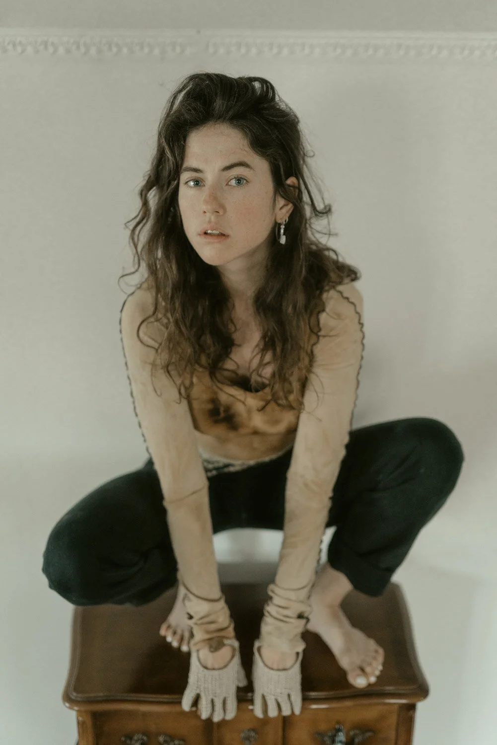Young woman with long, wavy brown hair and blue eyes sitting on a wooden table, wearing a beige long-sleeved top, black pants, knit fingerless gloves, and earrings, looking directly at the camera against a plain light-colored background.