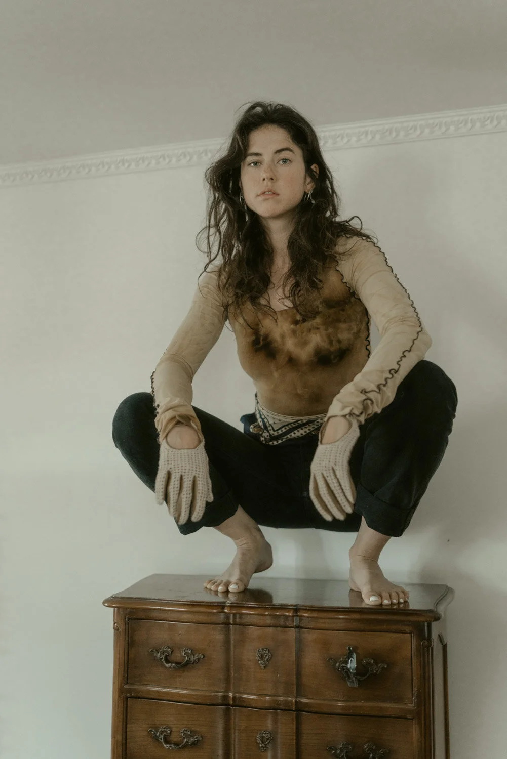 A young woman with curly brown hair balancing on a wooden dresser, wearing ripped black pants, a sheer beige top, and beige gloves with cutouts, squatting with her feet on the dresser and hands on her knees, looking at the camera.