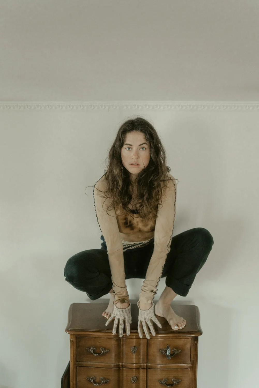 Woman with curly brown hair crouching on a wooden dresser, wearing beige gloves and no shoes, in a room with plain light-colored walls.