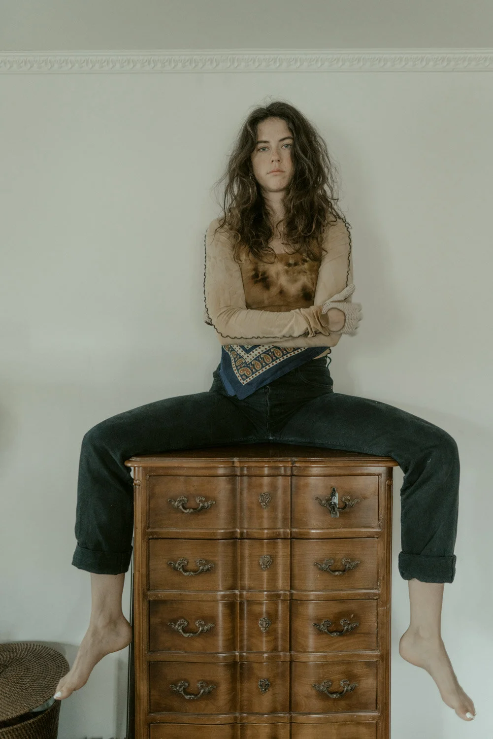 A young woman with long, curly hair sits cross-legged on a vintage wooden dresser, arms crossed, with a serious expression, against a plain wall.