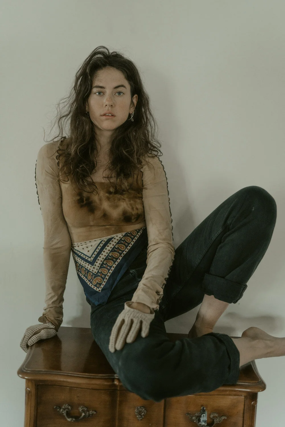 A woman with curly brown hair sitting on a wooden dresser, wearing a semi-transparent tan long-sleeve top, dark rolled-up jeans, and beige gloves, with a neutral expression, against a plain light-colored wall.