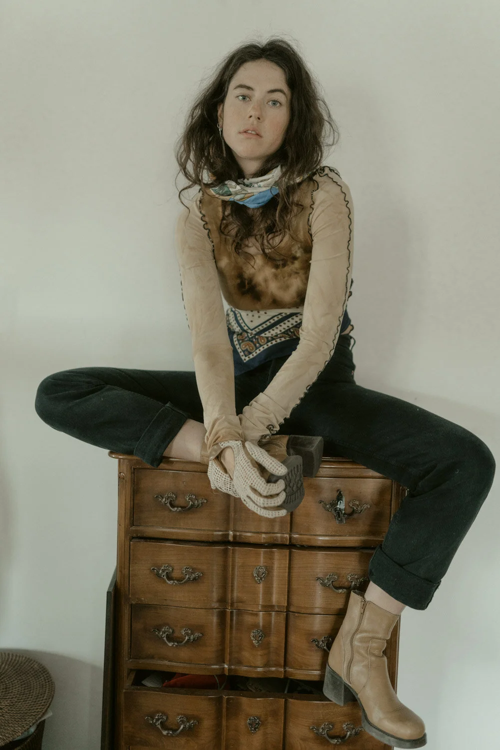 A woman sitting cross-legged on a wooden dresser, holding a hammer, wearing gloves, tan boots, and casual clothing, with a plain wall in the background.