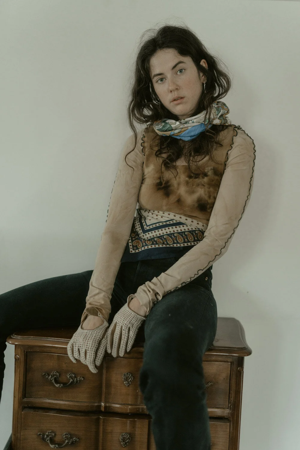 A young woman with dark curly hair wearing a colorful scarf, a patterned top, and black pants, sitting on a wooden dresser against a plain white wall.