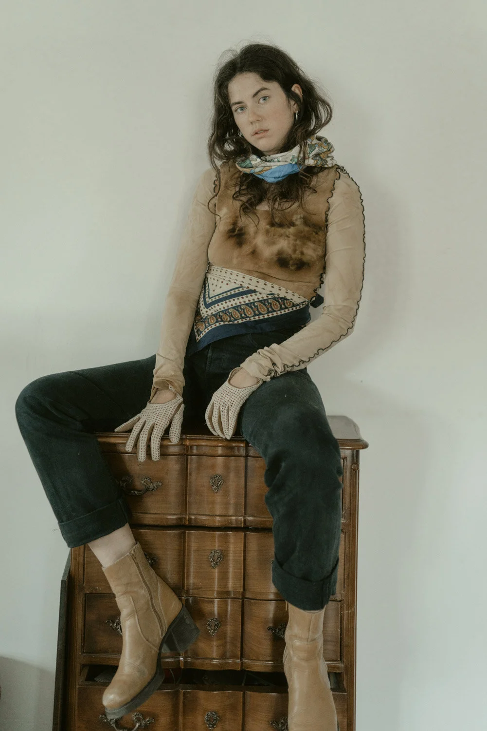 A young woman with dark, curly hair sitting on a vintage wooden dresser against a plain wall, wearing a sheer long-sleeve top, a colorful scarf, black pants, tan boots, and beige gloves.
