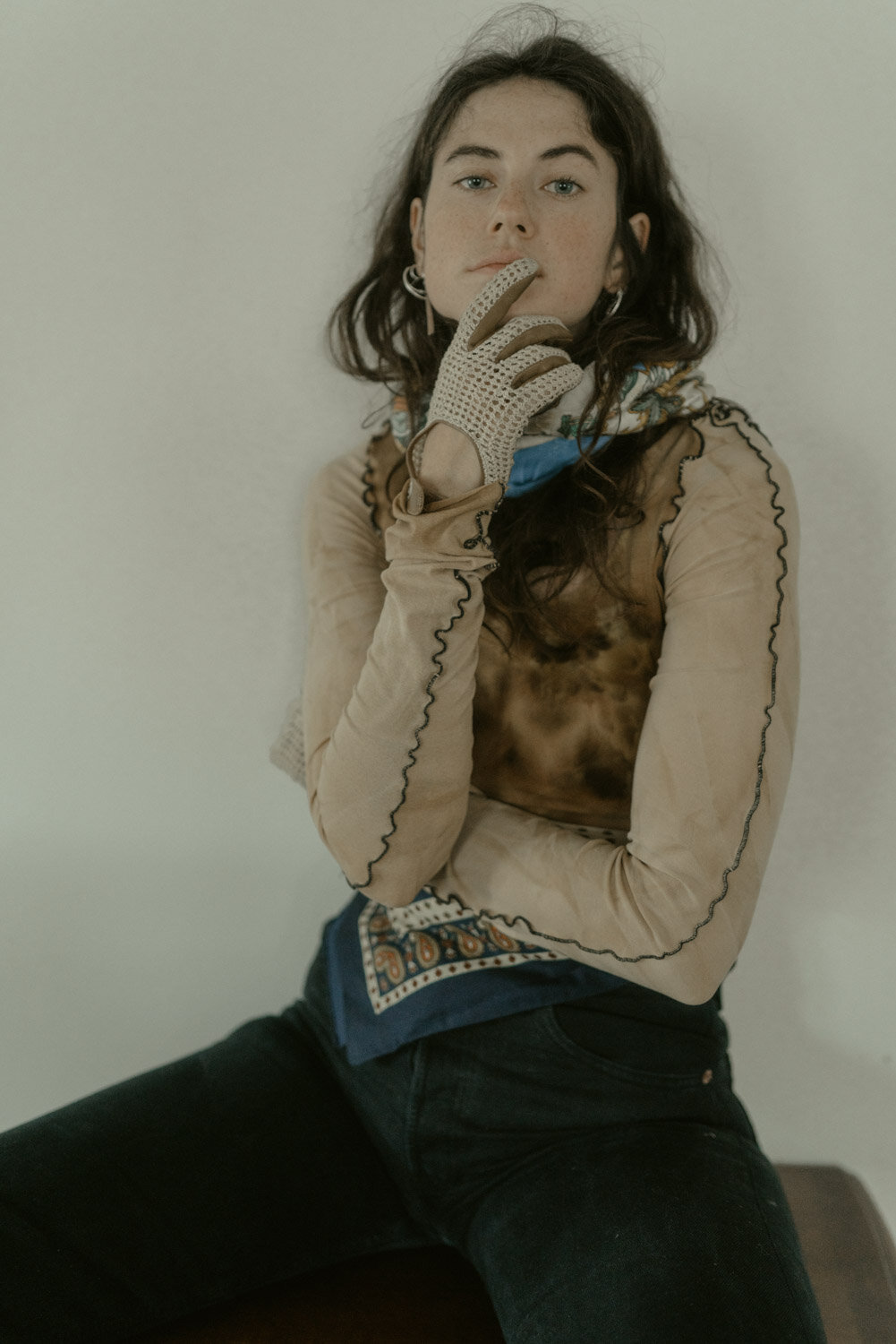 A young woman with dark, curly hair and fair skin posing against a plain wall, wearing a beige long-sleeve top with embroidered details, black jeans, and a patterned scarf, with a glove on her right hand resting near her lips.