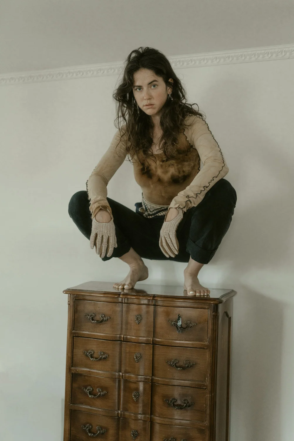 A woman squatting on top of a wooden dresser with a neutral expression, wearing beige gloves, a sheer long-sleeve shirt with a dark stain, and dark pants, with a plain white wall in the background.