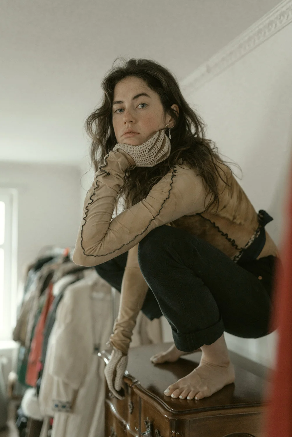 A young woman with dark, wavy hair squats on a wooden dresser, wearing a beige jacket, gloves, and dark jeans, looking directly at the camera with a serious expression.