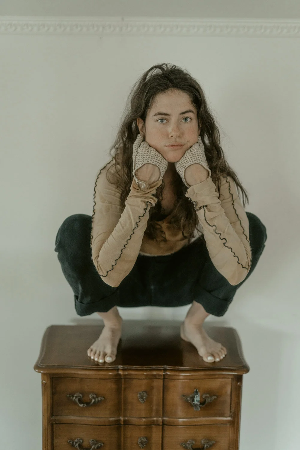 A woman with long dark curly hair squats on a vintage wooden dresser, resting her chin on her hands with gloves, looking directly at the camera with a neutral expression.