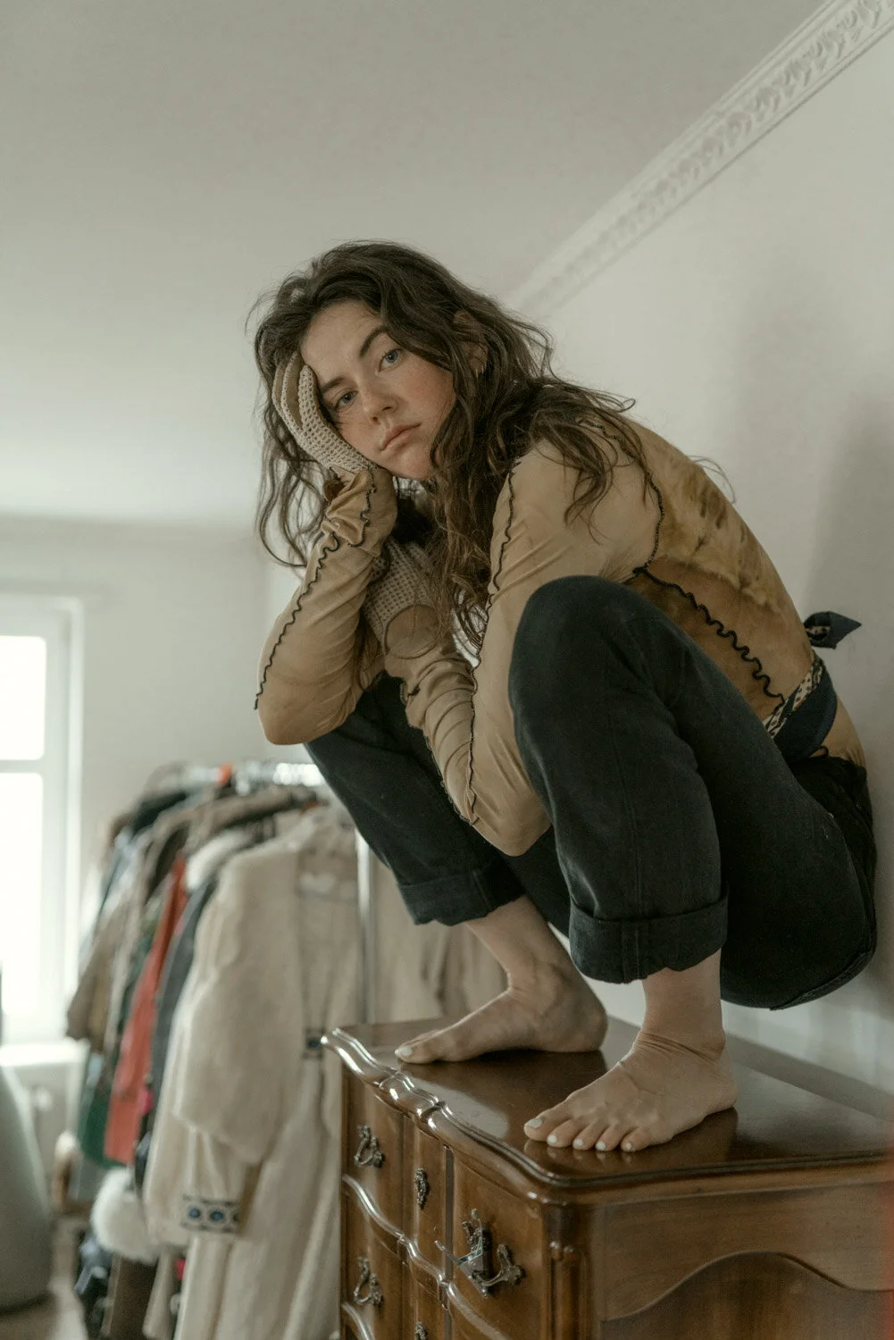 A woman with long, curly brown hair squats on a wooden dresser, her chin resting on her hand, looking into the distance with a pensive expression. She is wearing a beige, semi-transparent top with black stitching, black rolled-up pants, and beige glo