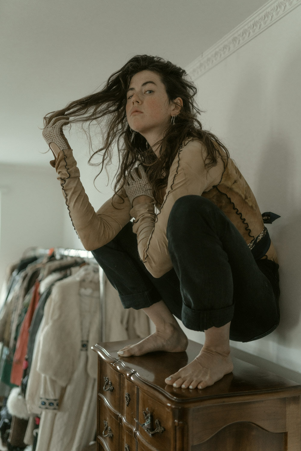 Woman squatting on a vintage wooden dresser, holding her hair with one hand and looking at the camera