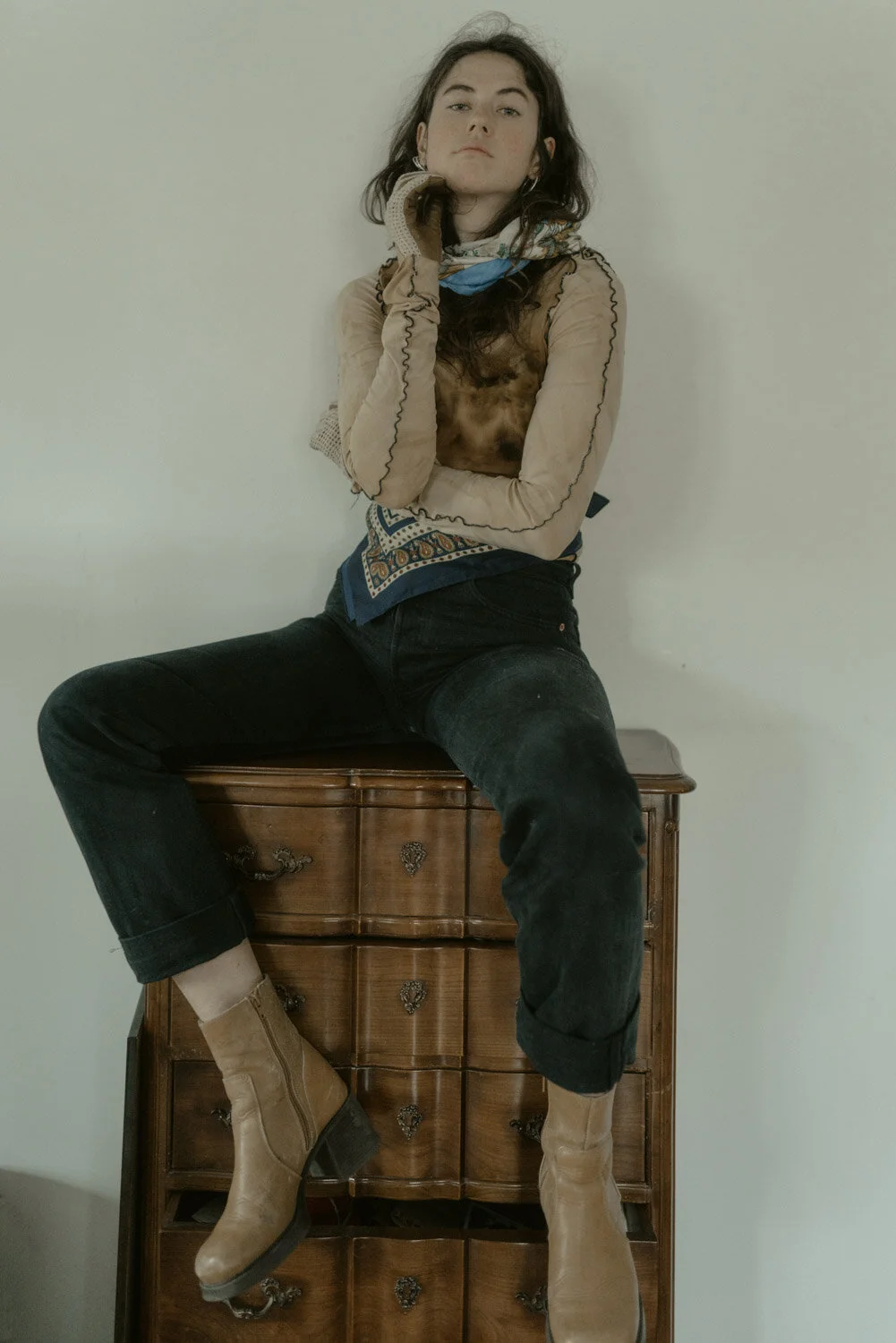 A young woman with dark hair and light skin sitting on a wooden dresser against a plain wall, wearing tan boots, black jeans, a layered top, and a colorful scarf.