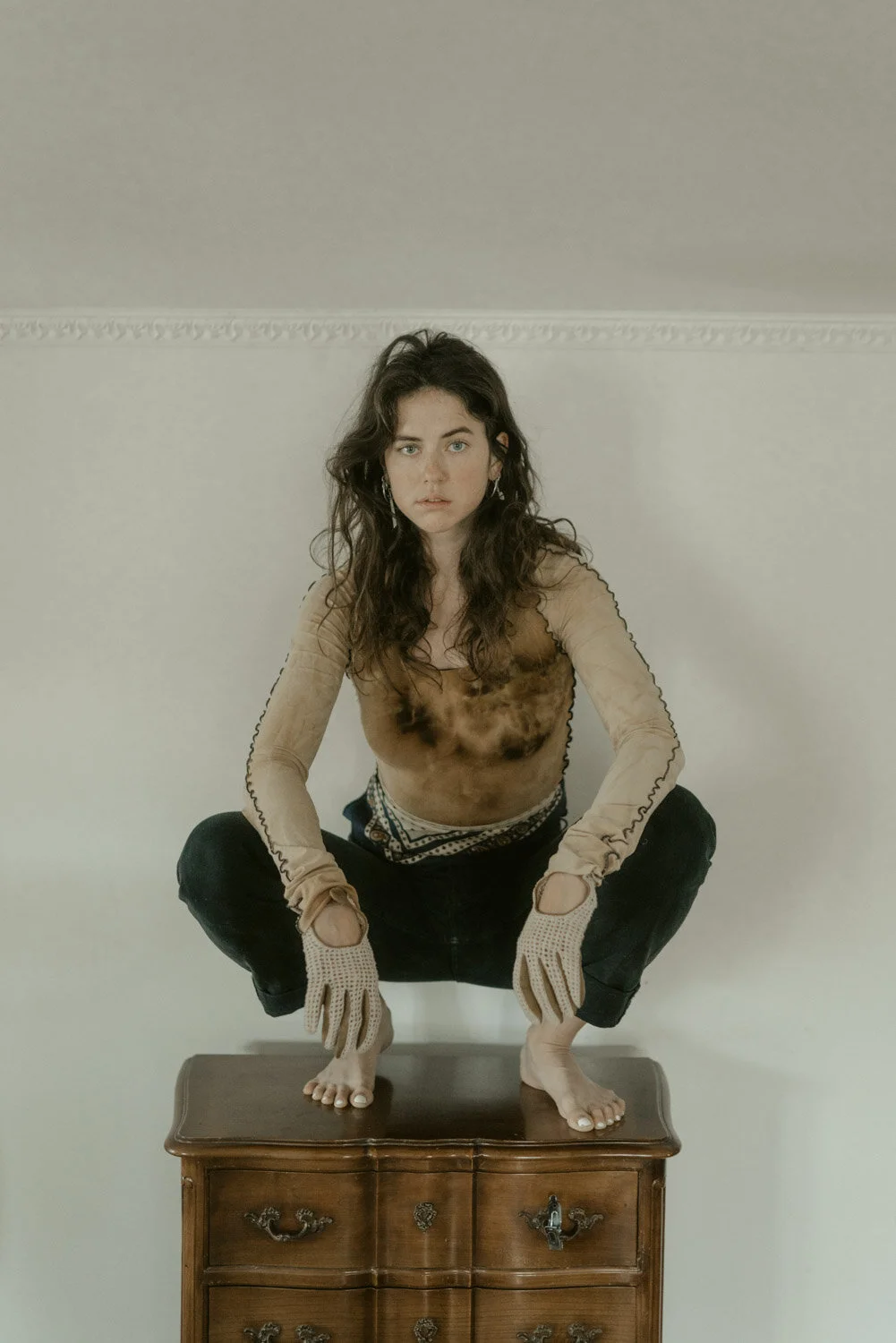 A woman with long dark hair squats on a wooden dresser against a plain white wall, wearing ripped black pants, beige gloves, and a sheer long-sleeve top with visible discoloration or stains.