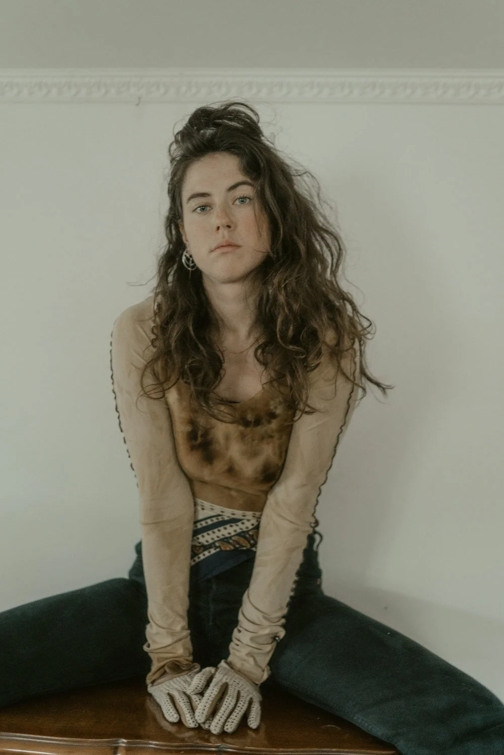 A young woman with long, curly brown hair sitting with legs apart on a wooden surface, leaning forward with her hands clasped together, wearing a beige long-sleeve top, patterned waistband, and gloves, against a plain white wall background.