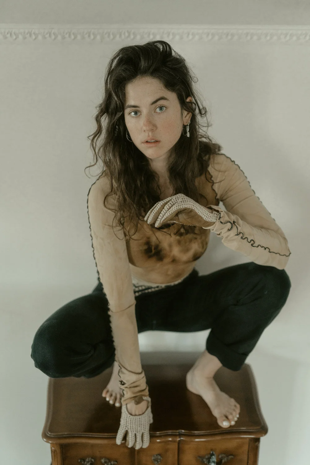 A woman with long wavy dark hair and blue eyes squats on a wooden table, wearing a sheer beige long-sleeve top with black accents, black pants, and a crochet glove on her right hand. She looks directly at the camera with a surprised expression agains