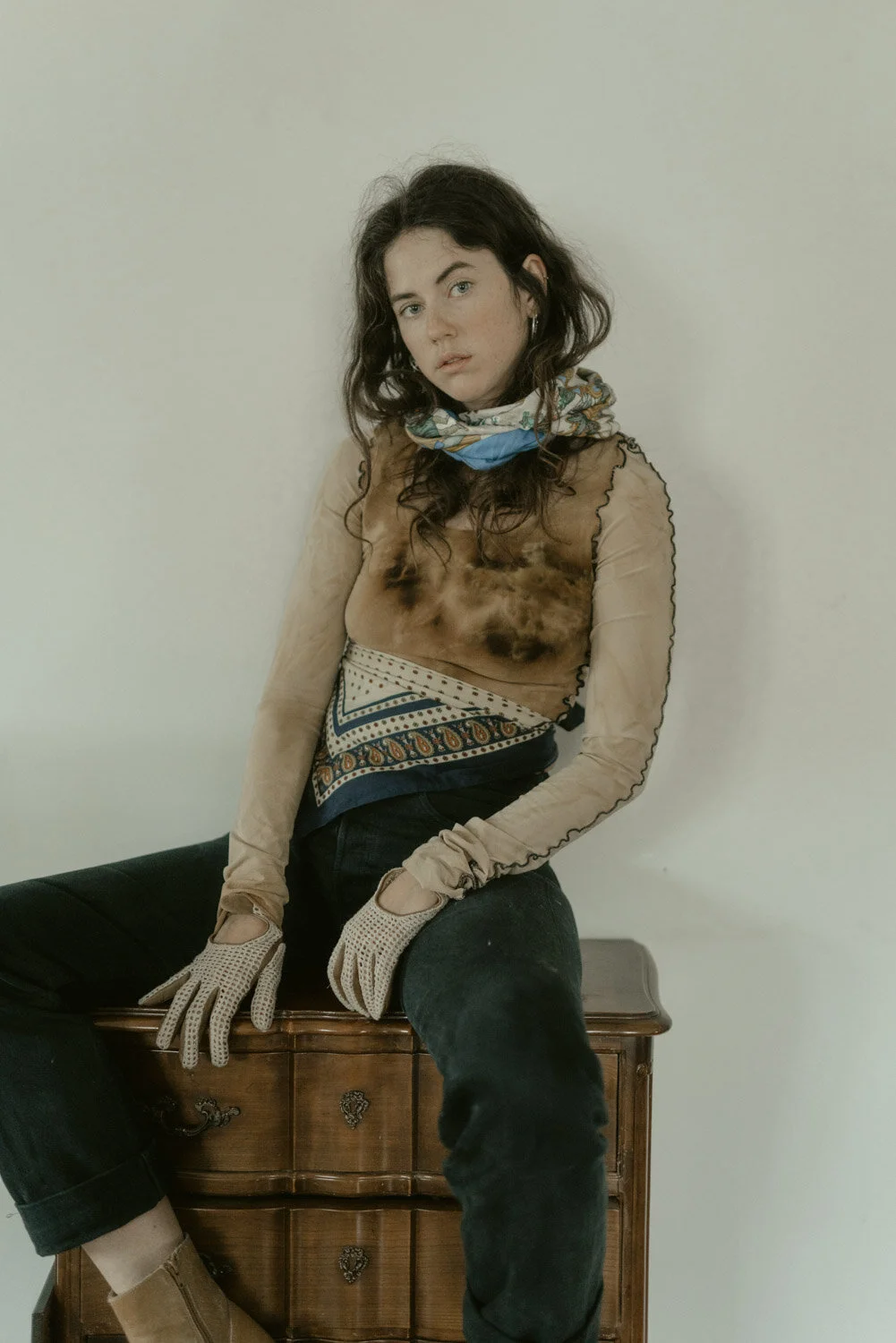 Young woman with dark curly hair sitting on a wooden chest, wearing gloves, a patterned scarf, sheer long-sleeve top, dark jeans, and beige boots, against a plain light-colored wall.