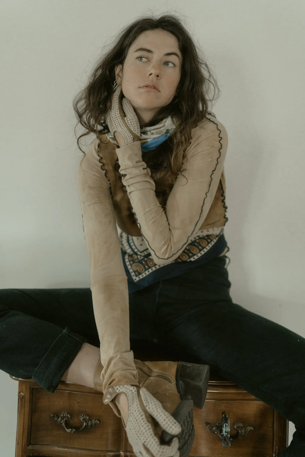 A young woman with wavy brown hair sits on a wooden dresser against a plain light-colored wall. She wears a tan long-sleeve top, patterned scarf, and black pants. She has a contemplative expression, with her right hand resting on her cheek and her le