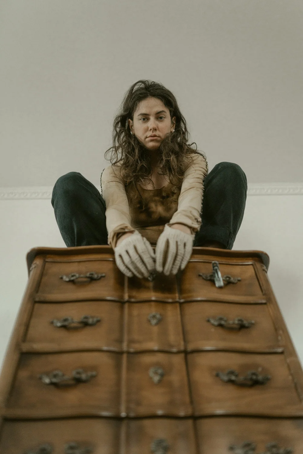 A young woman with curly dark hair, sitting cross-legged on a wooden dresser with multiple drawers, looking down at the camera with a serious expression, wearing beige gloves and a long-sleeved shirt.