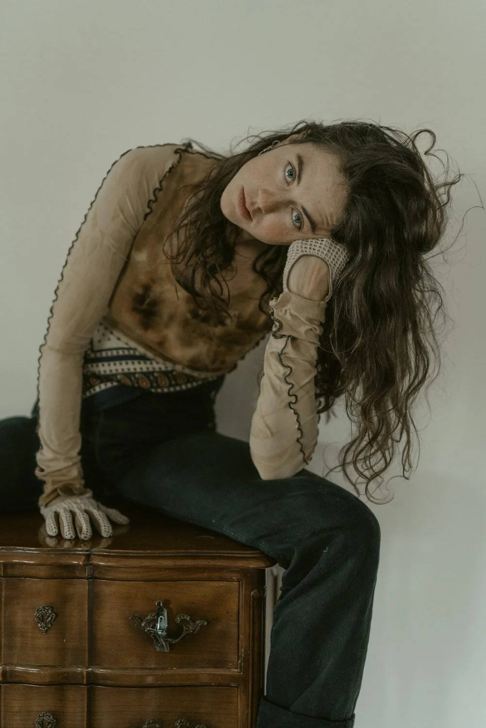 A woman with long wavy hair leaning on her hand while sitting on a wooden dresser, wearing a patterned shirt, black pants, and gloves, with a neutral background.