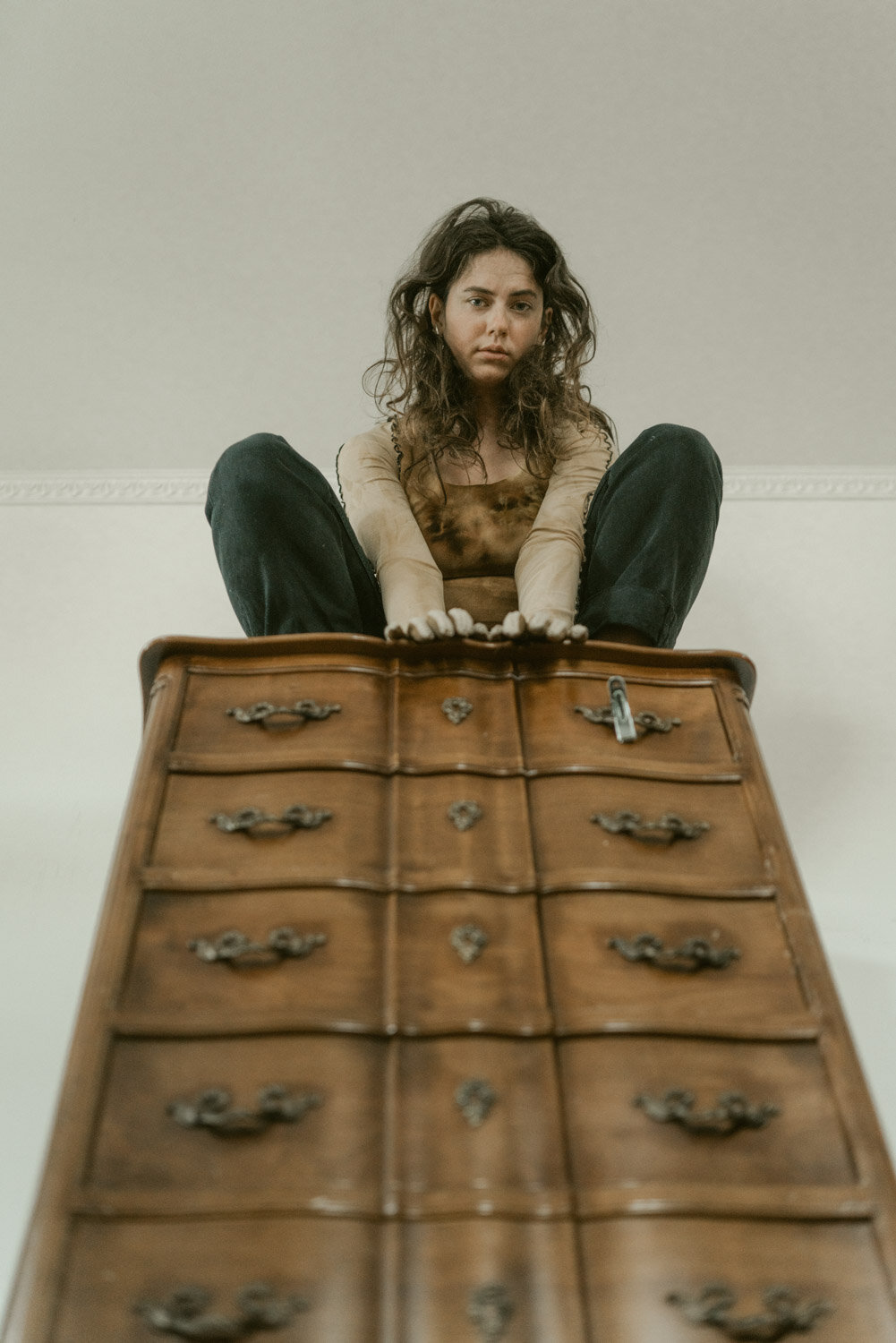 A woman with curly hair sitting on top of a wooden dresser, looking down at the camera with a serious expression.