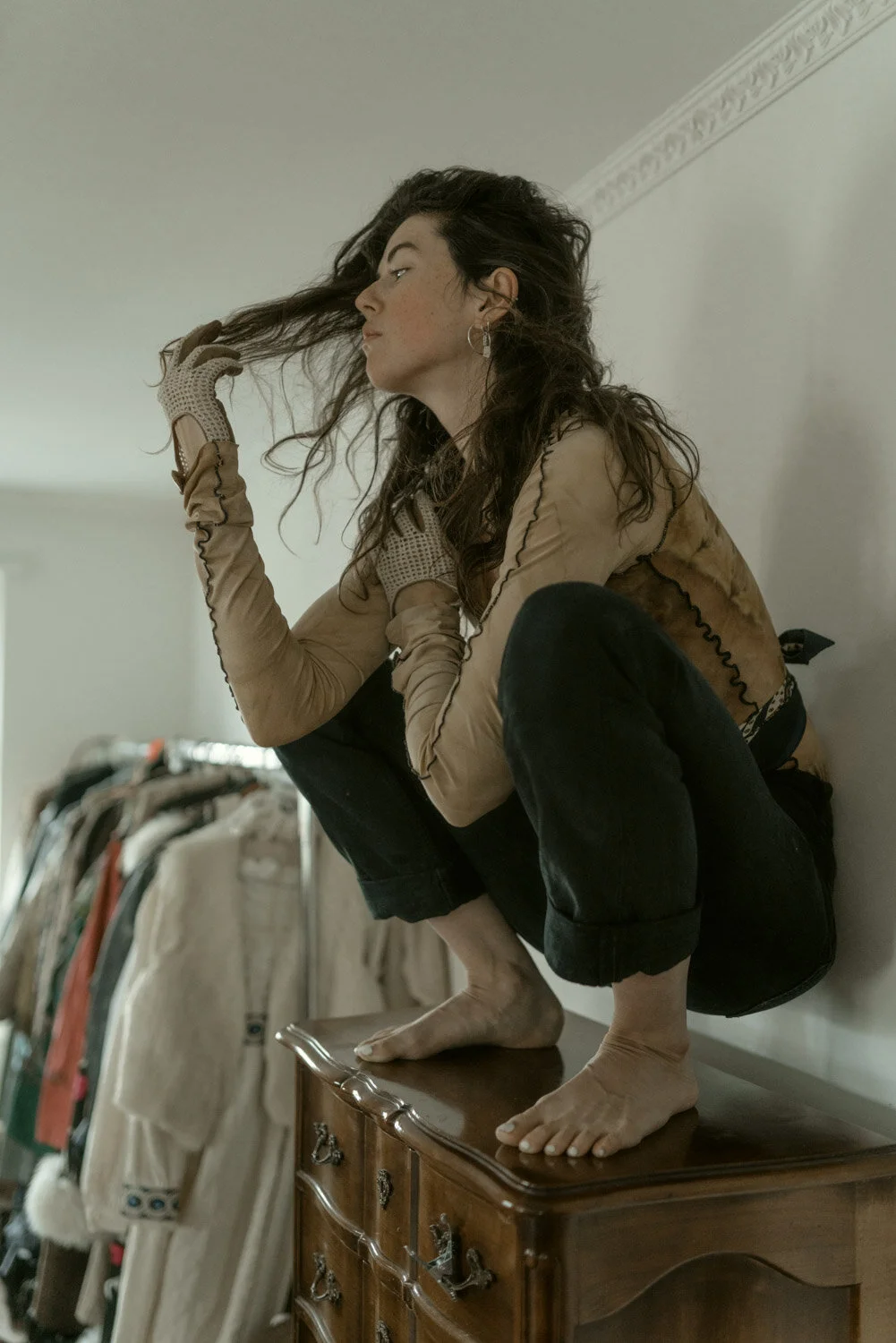 A woman with curly dark hair squats on a vintage wooden dresser, barefoot, wearing beige gloves and a beige jacket, gazing sideways and holding her hair with one hand.