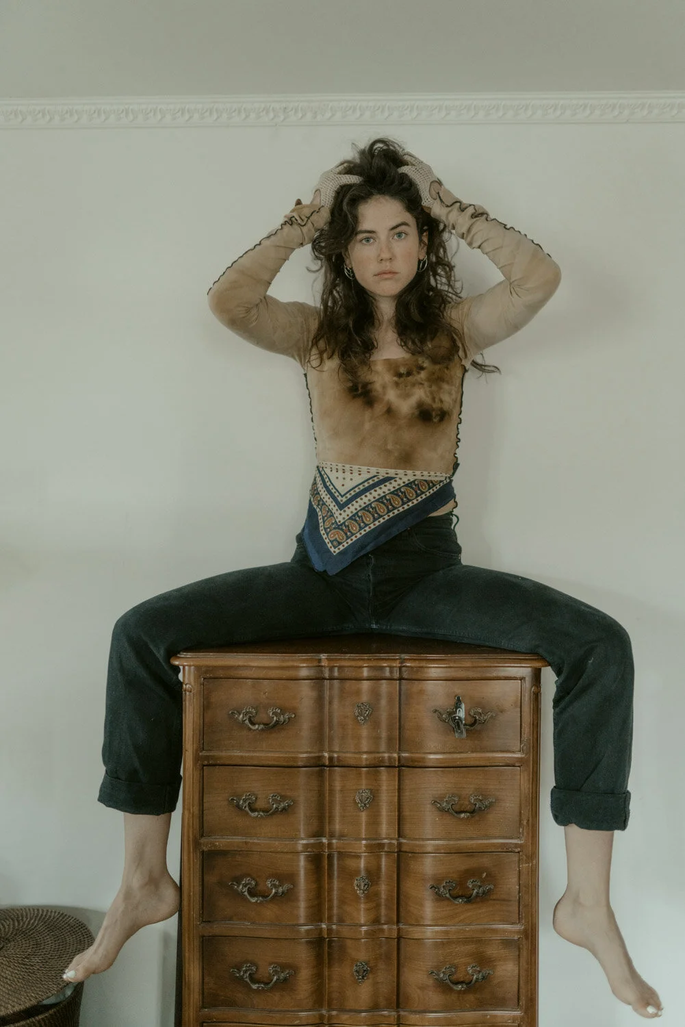 A woman with long curly hair is sitting on a wooden dresser with her legs spread wide apart, wearing a sheer long-sleeve top, a bandana around her waist, and black jeans, with her arms raised above her head and hands in her hair.