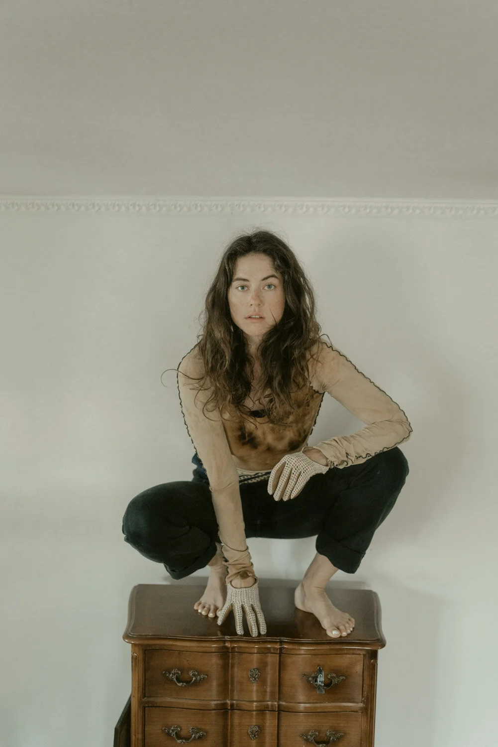 A woman with long, curly brown hair and blue eyes crouches on top of a wooden dresser, looking directly at the camera. She is wearing a beige long-sleeve top, black pants, and white gloves. The background is a plain, light-colored wall.