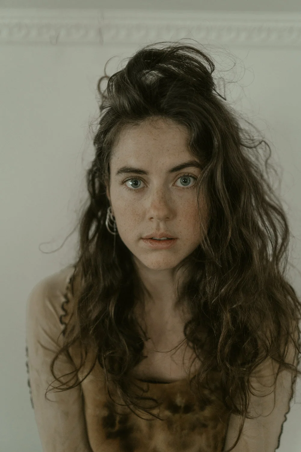 A young woman with blue eyes, freckled face, and long, curly brown hair, wearing a beige tie-dye shirt and hoop earrings.