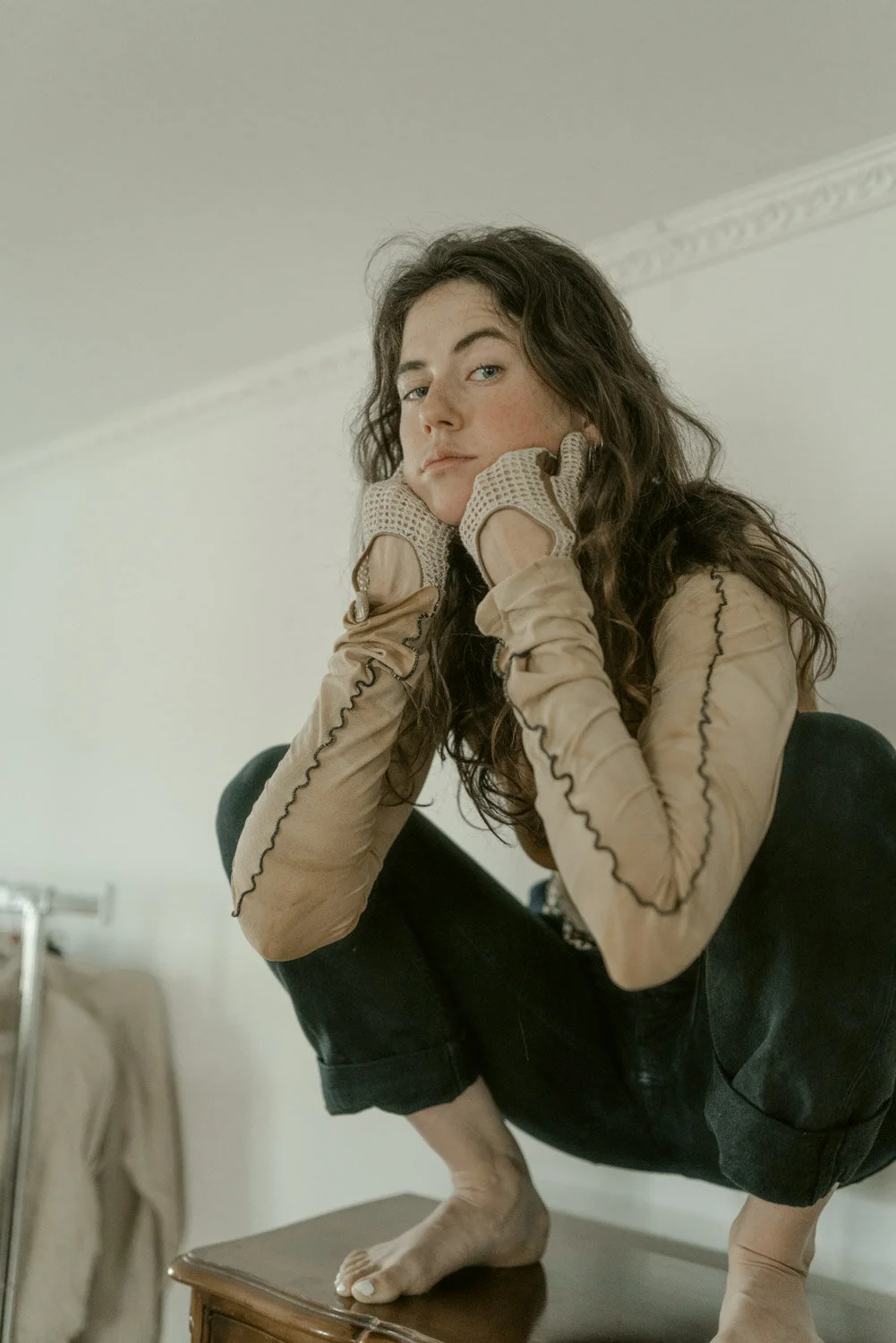 A woman with curly hair squats on a table, resting her chin on her hands, wearing beige gloves and a beige top with black stitching details.