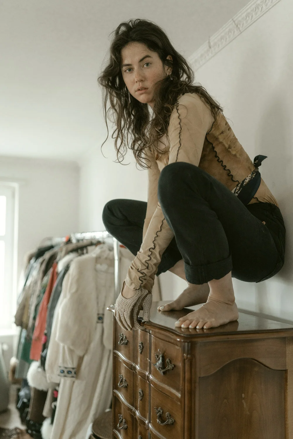 Woman crouching on a wooden dresser, wearing a beige top, black jeans, and gloves, in a room with clothing hanging on racks.