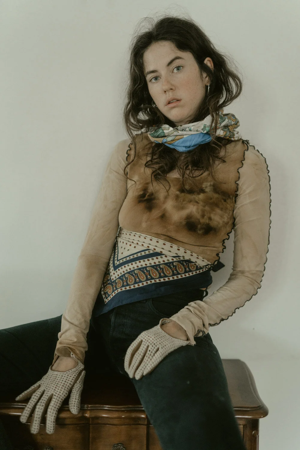 Young woman with dark, curly hair, wearing a patterned scarf, sheer long-sleeve shirt, and black pants, sitting on a wooden table, looking at the camera.