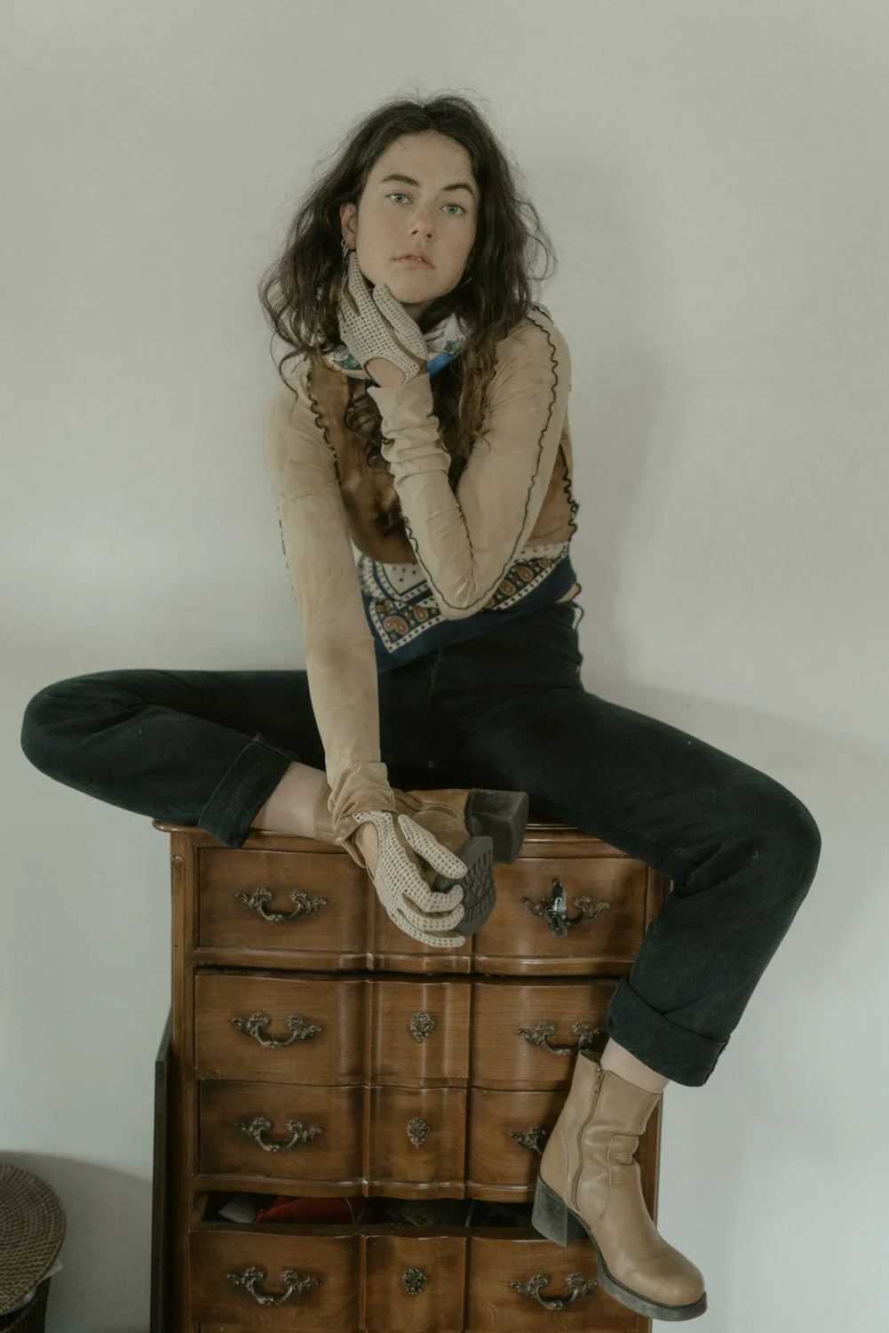 A woman with long, curly dark hair sitting on a vintage wooden dresser, wearing beige boots, black pants, a patterned shirt, a leather vest, and beige gloves, posing with her hand on her chin.