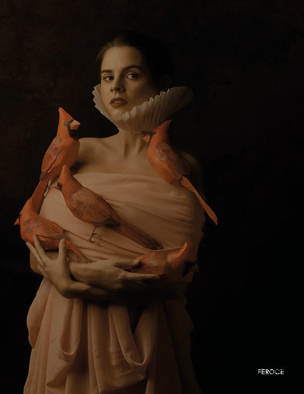 A woman with dark hair and makeup wearing an elaborate beige dress with large ruffles, surrounded by orange birds perched on her arms and shoulders, against a dark background.