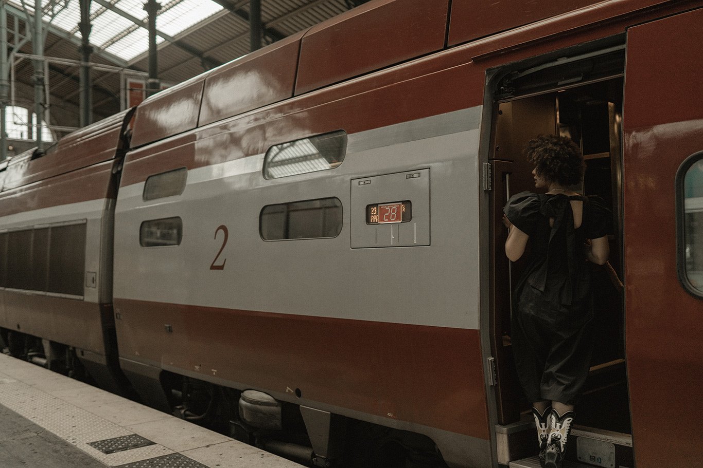 A person with curly hair wearing a black dress and patterned boots is boarding a red and white train at a station.