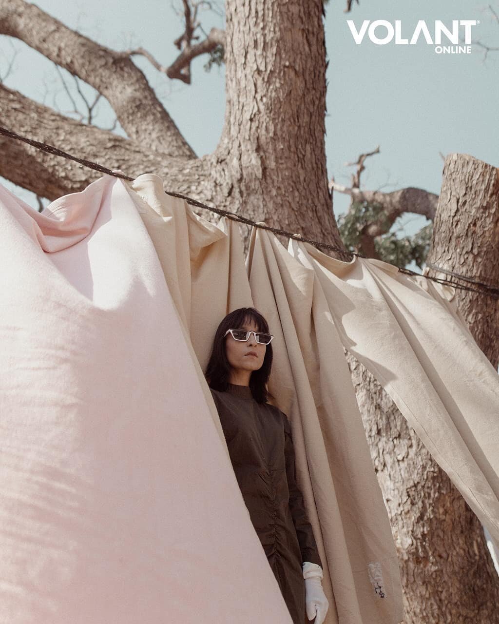 A woman with dark hair, wearing sunglasses, a brown dress, white gloves, standing among beige curtains hanging from a tree branch outdoors.
