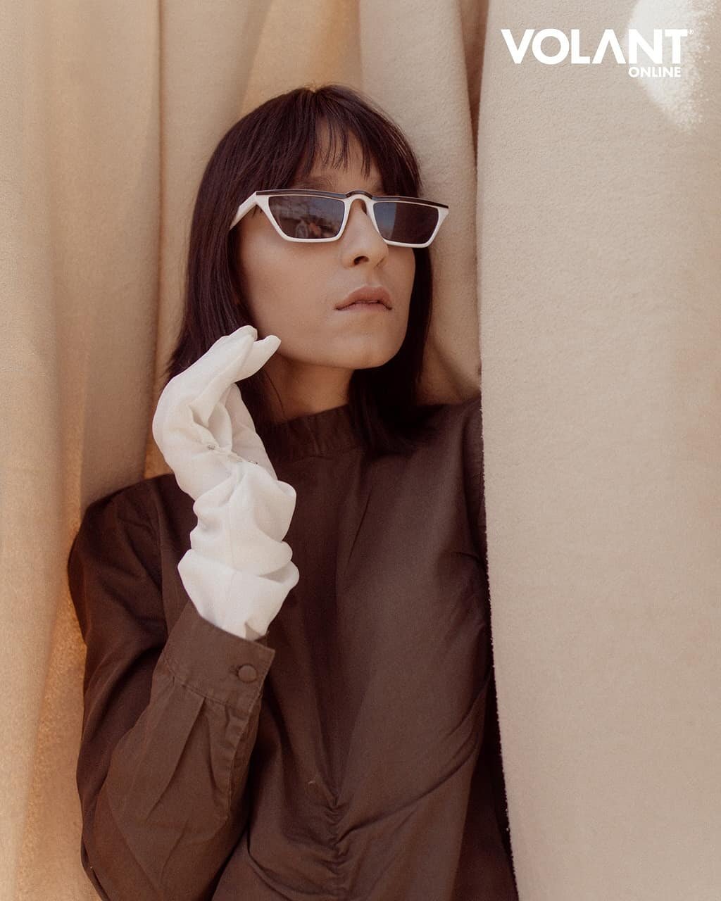 A woman wearing sunglasses and a brown shirt with a white glove on her left hand, posed against beige curtains.