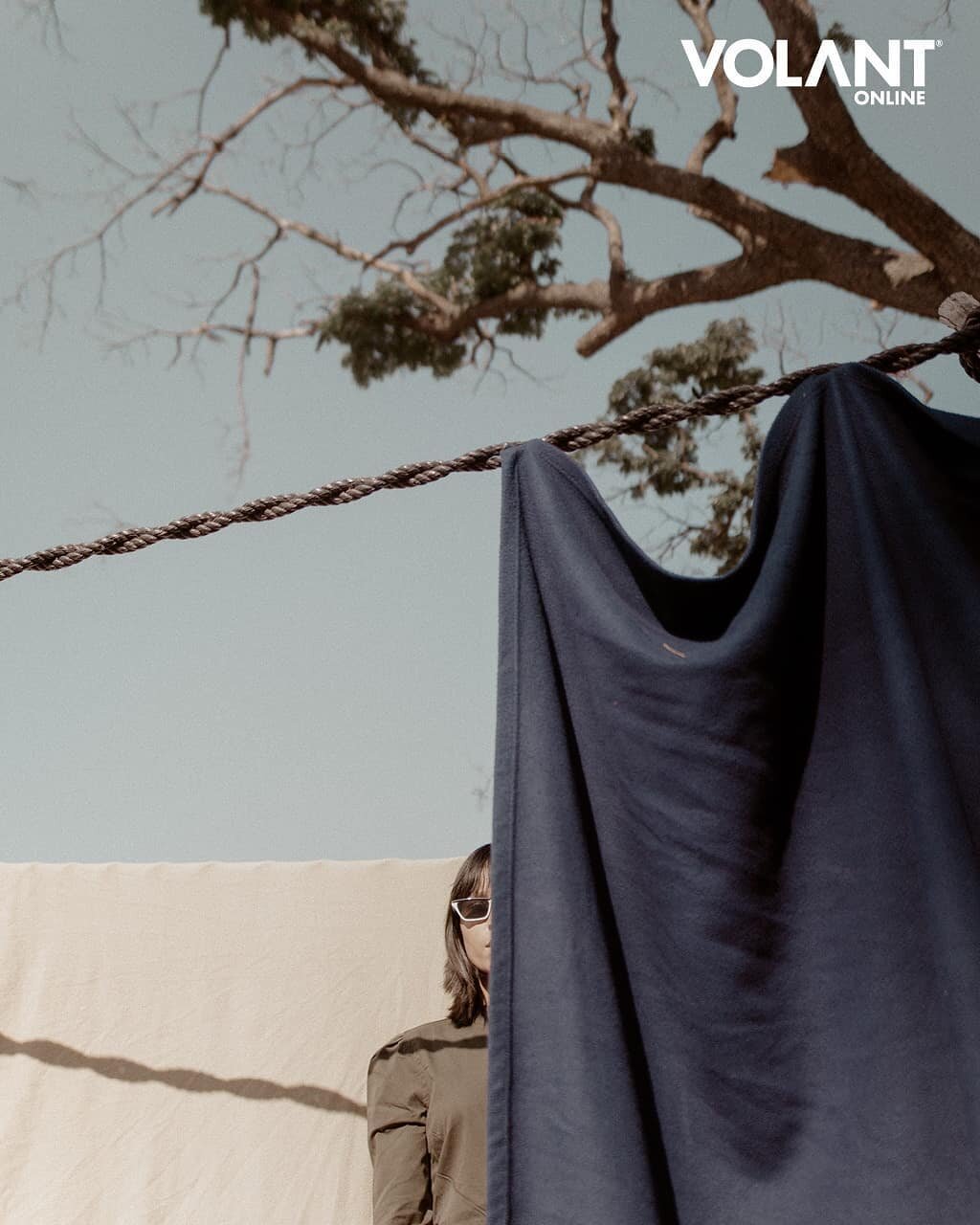 A person with brown hair and sunglasses partially hidden behind a hanging black cloth outdoors. String is tied to a tree branch in the background. The sky is overcast, and the scene appears to be a casual outdoor setting.
