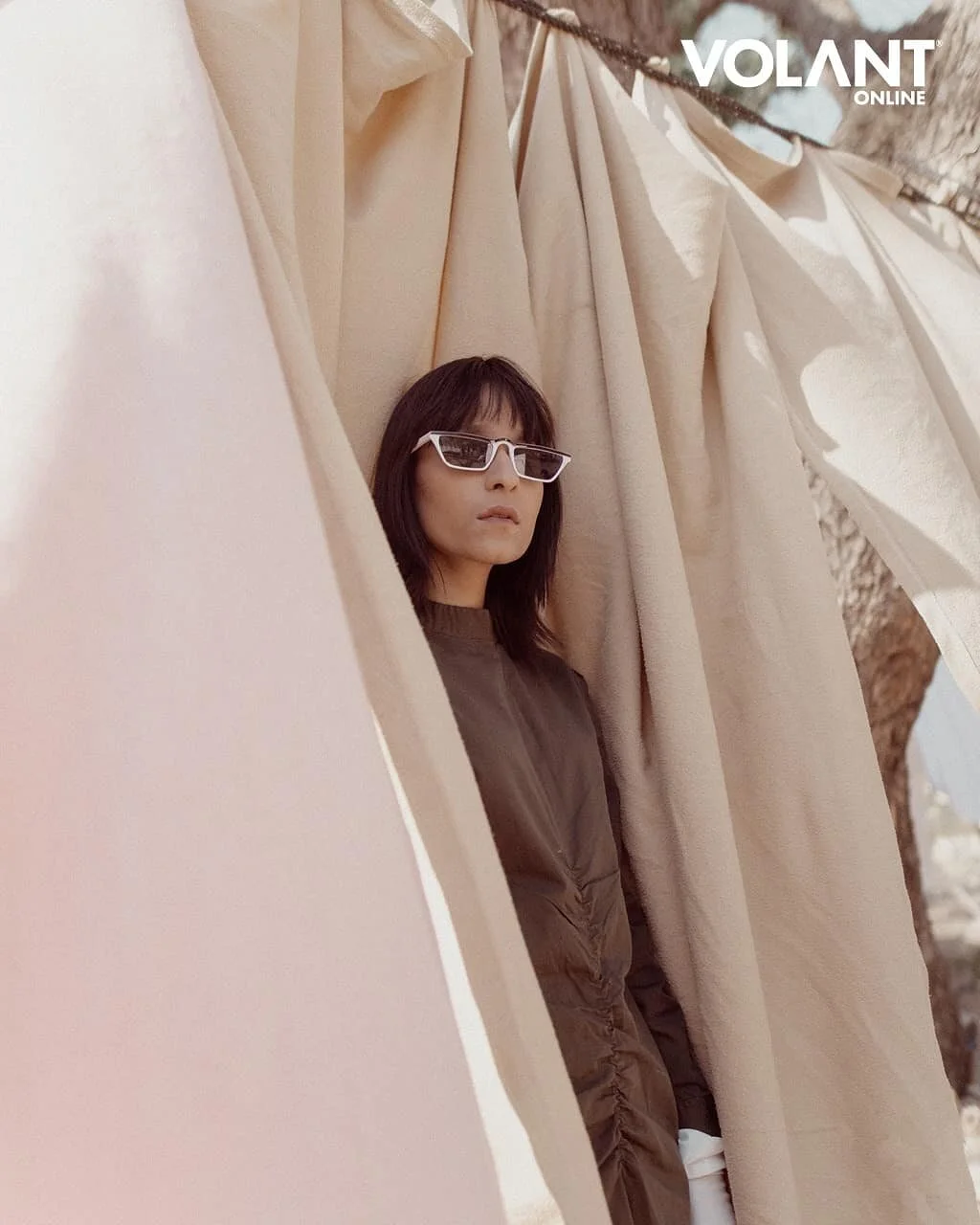 A woman with shoulder-length dark hair wearing white sunglasses and a brown jacket, standing among beige curtains outdoors.