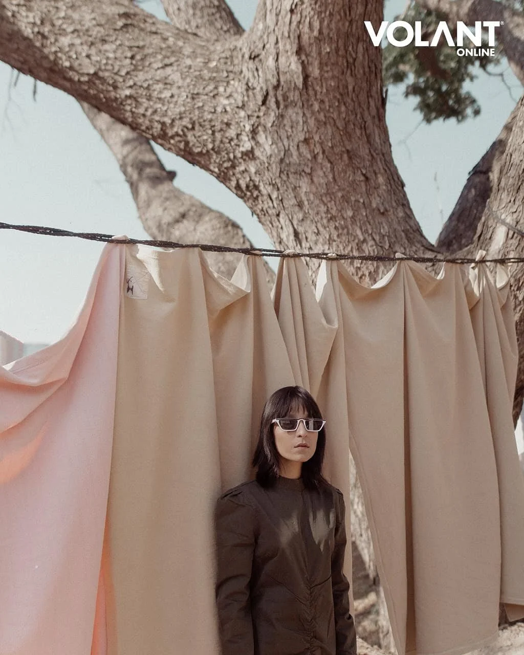 A woman with dark hair and sunglasses standing behind beige curtains suspended from a line, with a large tree trunk and branches in the background.
