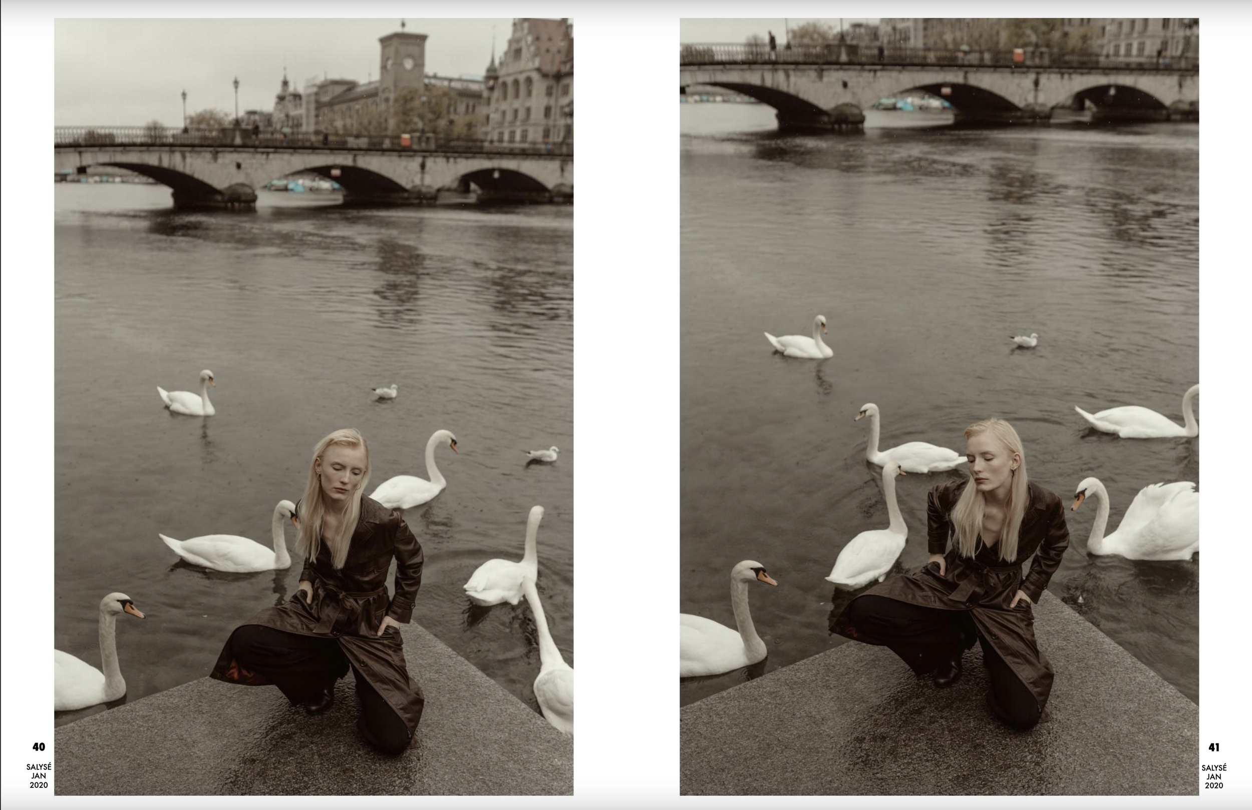 A woman kneeling on the ground near a river, surrounded by swans, with a bridge and cityscape in the background.