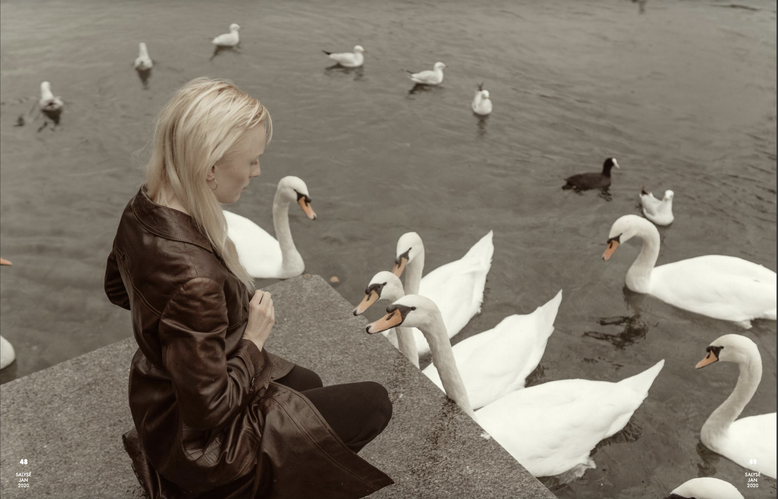 A woman in a brown leather jacket sitting by the water's edge, feeding or interacting with a group of swans on a gray, overcast day.