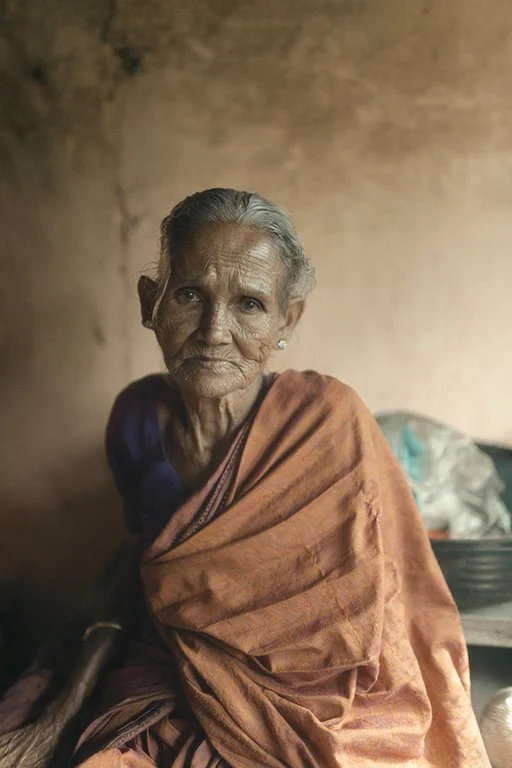 An elderly woman with gray hair, wearing a dark top and an orange shawl, sitting indoors against a plain wall.