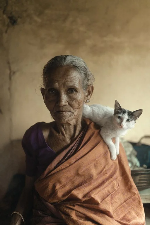 An elderly woman with gray hair and a serious expression, wearing an orange traditional sari, is holding a white and gray cat on her shoulder in a dimly lit room with a plain, slightly textured wall in the background.